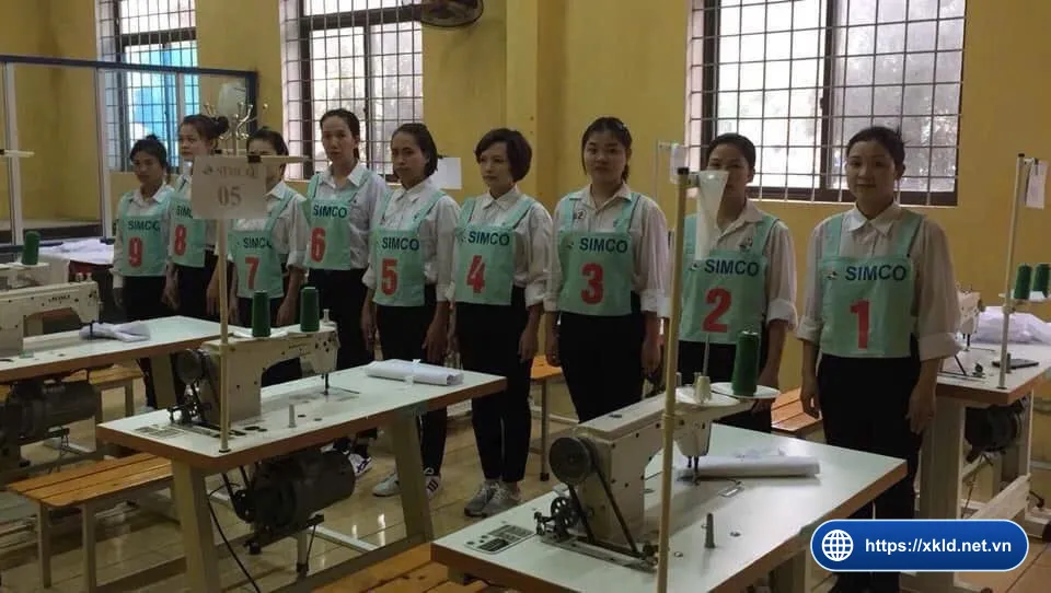 Chín lao động nữ mặc áo sơ mi trắng, đeo yếm xanh ghi SIMCO số 1 đến 9, đứng thành hàng cạnh các bàn máy may công nghiệp trong phòng học có cửa sổ, trong buổi thi tuyển đơn hàng may mặc của SIMCO Sông Đà