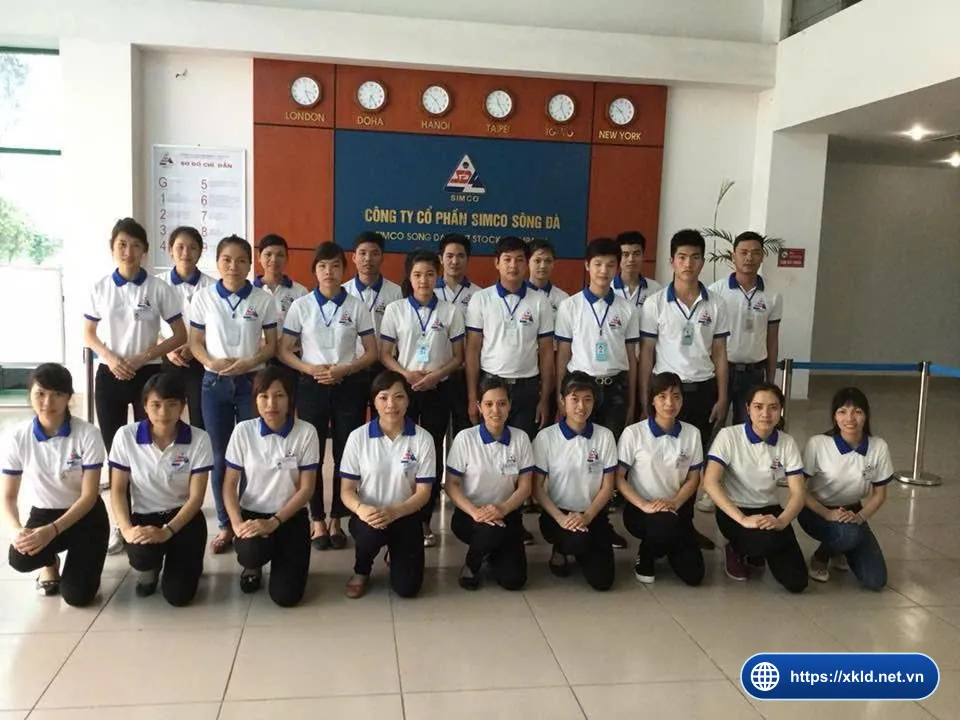 Nhóm lao động nam và nữ mặc áo thun trắng viền xanh, hàng sau đứng, hàng trước quỳ gối, chụp trước biển Công ty Cổ phần SIMCO Sông Đà và dãy đồng hồ các thành phố