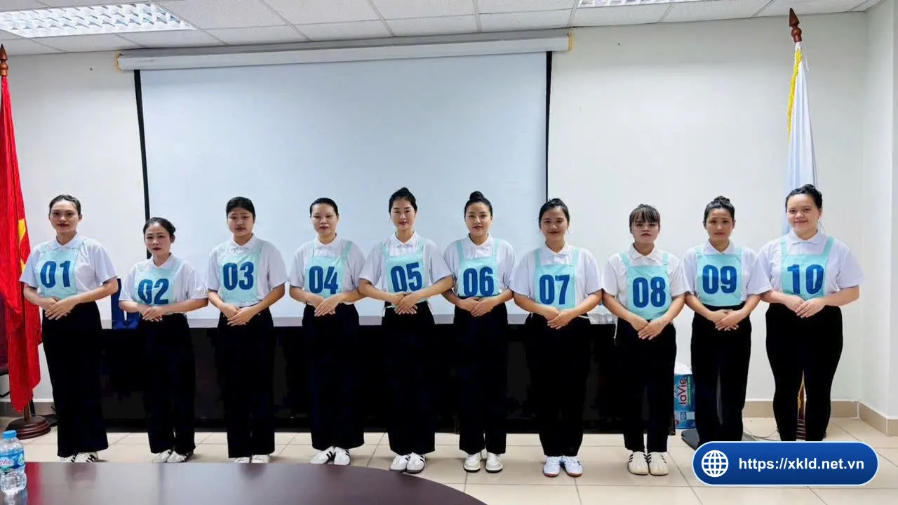 Mười lao động nữ mặc áo sơ mi trắng, đeo số báo danh từ 01 đến 10, đứng thành hàng trước màn chiếu và lá cờ đỏ trong phòng họp của văn phòng SIMCO Sông Đà