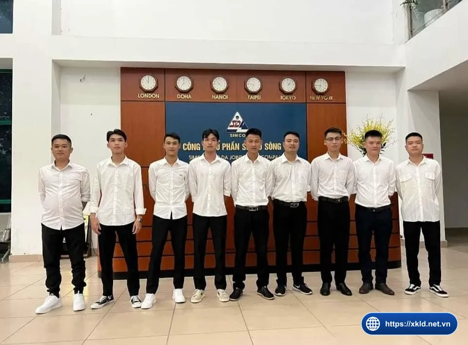 Chín lao động nam mặc áo sơ mi trắng, quần đen, đứng thành hàng ngang trước quầy lễ tân có logo SIMCO Sông Đà và dãy đồng hồ các thành phố