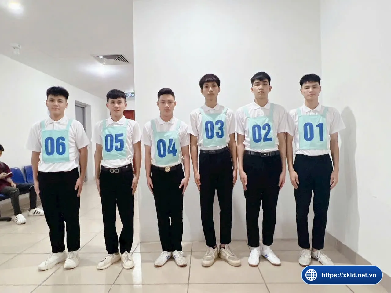 Sáu lao động nam mặc áo sơ mi trắng, quần đen, đeo số từ 01 đến 06 đứng thành hàng dọc theo bức tường trong phòng chờ thi tuyển đơn hàng của SIMCO Sông Đà