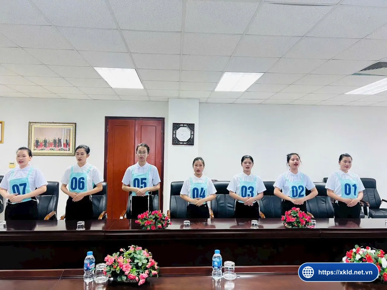 Bảy lao động nữ mặc áo sơ mi trắng, đeo số báo danh từ 01 đến 07, đứng thành hàng sau bàn họp dài có hoa tươi trong một phòng họp của văn phòng SIMCO Sông Đà
