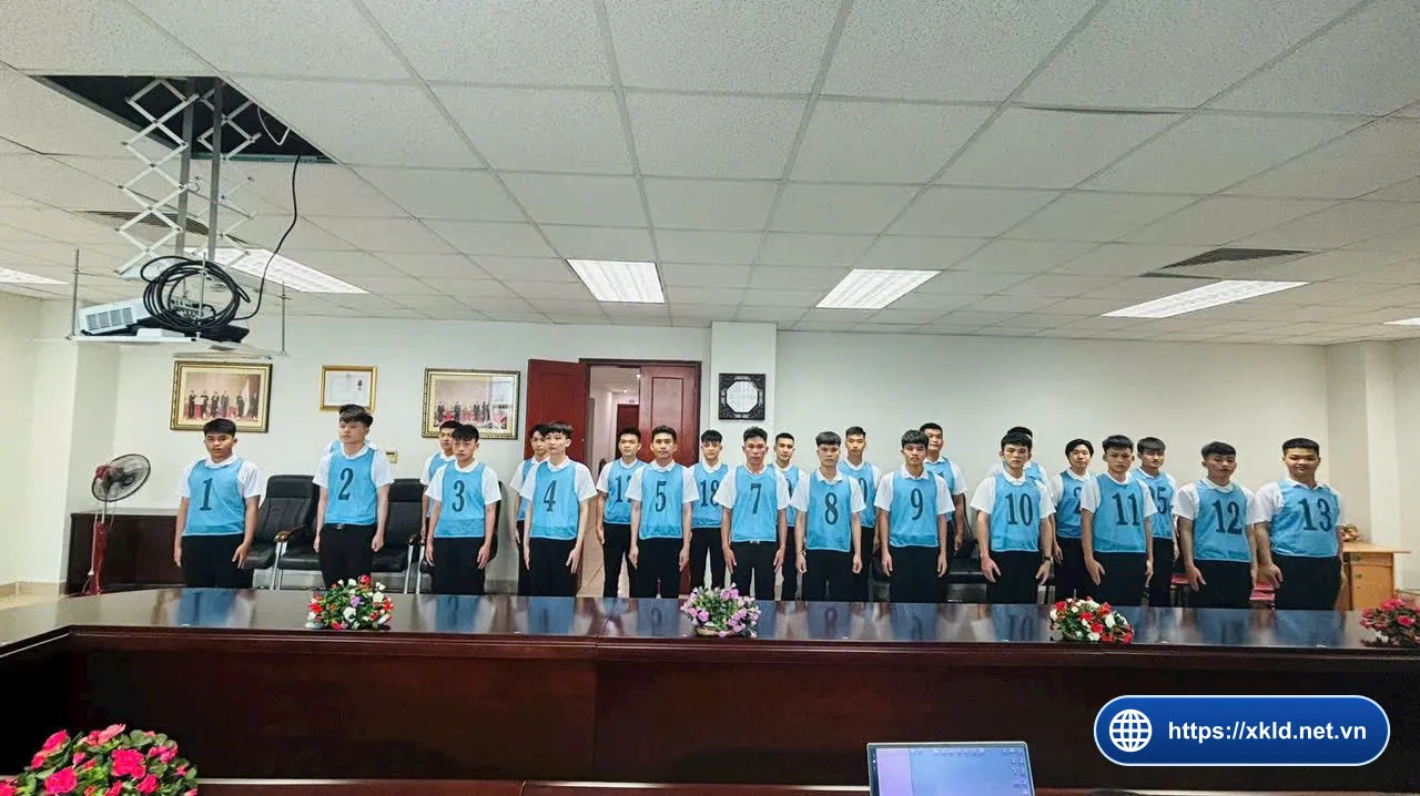 Nhóm lao động nam mặc áo thun trắng, đeo yếm xanh dương đánh số từ 1 đến 13 trở lên, đứng thành hàng sau bàn họp dài trong phòng có hoa tươi, chờ đến lượt thi tuyển đơn hàng của SIMCO Sông Đà