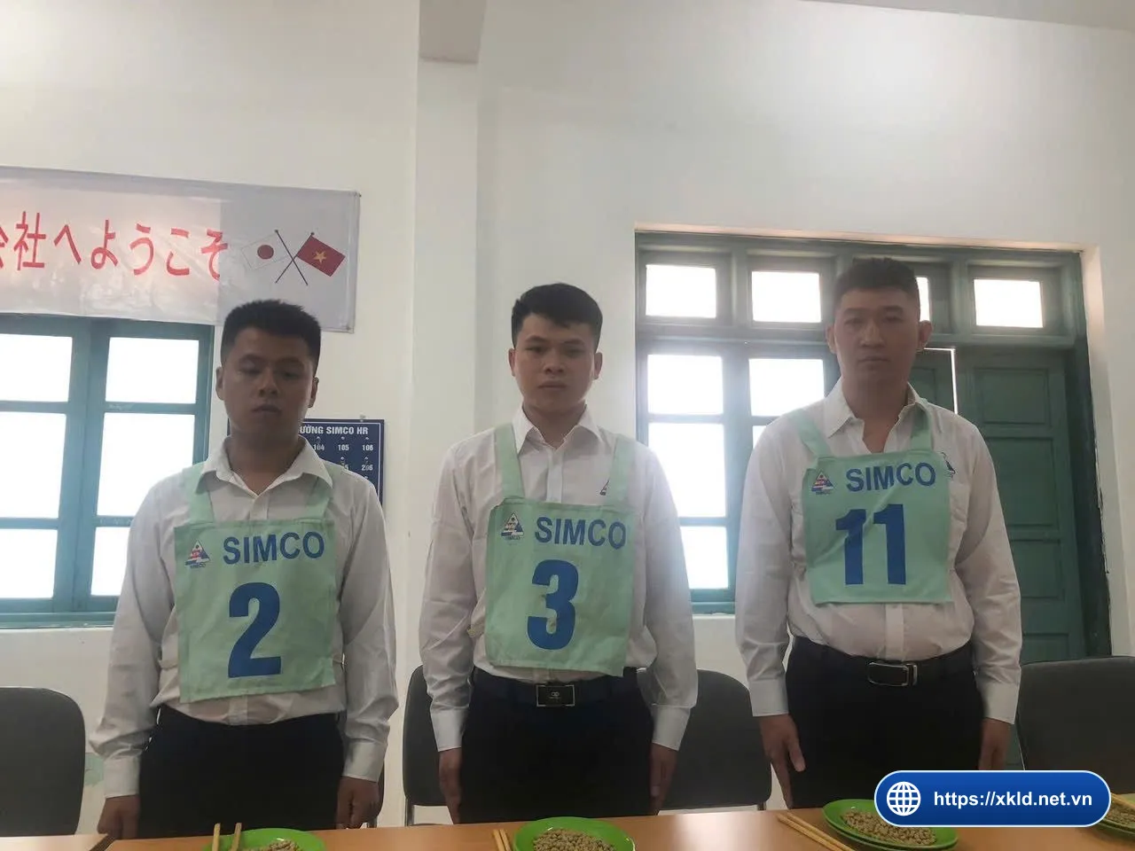 Ba lao động nam mặc áo sơ mi trắng, đeo yếm xanh ghi SIMCO số 2, 3 và 11, đứng sau bàn có đĩa xanh và đũa, phía sau là băng rôn tiếng Nhật với cờ Nhật Bản và cờ Việt Nam, trong buổi thi tuyển đơn hàng của SIMCO Sông Đà