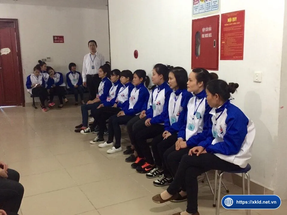 Nhóm lao động nữ mặc áo khoác xanh trắng đeo thẻ số ngồi thành hàng trên ghế trong phòng chờ, một cán bộ nam áo sơ mi trắng đứng phía sau, chờ đến lượt phỏng vấn đơn hàng của SIMCO Sông Đà